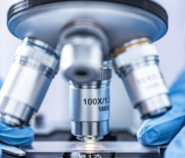 Microscope lenses with hands in blue rubber gloves holding a sample underneath..